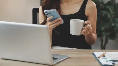 Genç Asyalı kadın dizüstü bilgisayarla çalışıyor ve elinde kahve tutuyor.