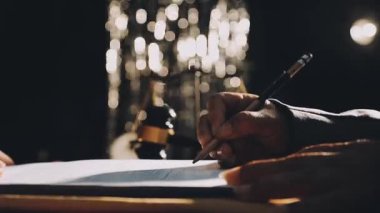 Karanlık bir arkaplan önünde siyah bir masanın üzerine kalemle yazan bir kişi.