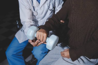 Evde dambıl kullanarak fizyoterapist eğitimi alan yaşlı bir kadın. Terapist, huzurevi egzersizlerinde yaşlı kadına yardım ediyor. Yaşlı bir hasta kollarını uzatmış halterler kullanıyor..