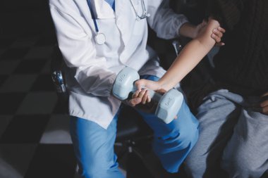 Evde dambıl kullanarak fizyoterapist eğitimi alan yaşlı bir kadın. Terapist, huzurevi egzersizlerinde yaşlı kadına yardım ediyor. Yaşlı bir hasta kollarını uzatmış halterler kullanıyor..