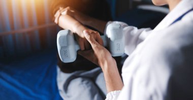 Evde dambıl kullanarak fizyoterapist eğitimi alan yaşlı bir kadın. Terapist, huzurevi egzersizlerinde yaşlı kadına yardım ediyor. Yaşlı bir hasta kollarını uzatmış halterler kullanıyor..