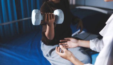 Evde dambıl kullanarak fizyoterapist eğitimi alan yaşlı bir kadın. Terapist, huzurevi egzersizlerinde yaşlı kadına yardım ediyor. Yaşlı bir hasta kollarını uzatmış halterler kullanıyor..