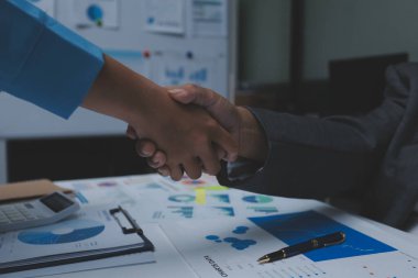 Girişimcilerin işbirliği anlaşması modern bir ofiste el sıkışıyor ve masadaki finansal kağıt grafiği.