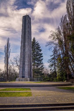 Üzerinde haç bulunan yüksek dikdörtgen beton bir anıt kulesi çıplak ağaçlarla çevrili bir parkta dramatik bulutlu bir gökyüzünün altında..