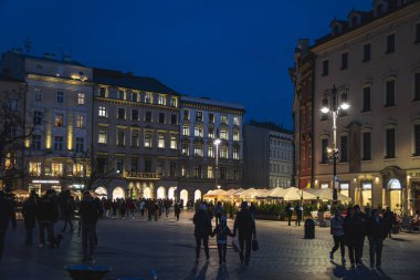 Rynek Glowny (Main Market Square) in Krakow's Old Town at twilight, showing historic townhouses with illuminated facades, arcade colonnade, market stalls, and evening crowds on cobblestone plaza