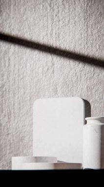 Beyaz mermer podyumlar ahşap masanın üzerinde ürün sergilenmesi için. Vazo ve desenli duvarlı minimalist 3D işlemeli. Doğal ışıklandırma ve güçlü gölgelerle kozmetik ya da cilt bakımı için günışığı arka planı.