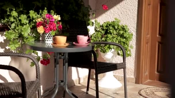 Tasses orange et rose avec café ou thé sur la table sur le porche de la maison entre les fleurs. — Video