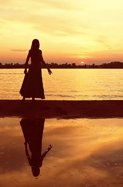 Una ragazza in abito lungo si erge su uno spiedo di sabbia riflesso nelle acque del fiume e guarda il sole che tramonta all'orizzonte. Immagine Stock