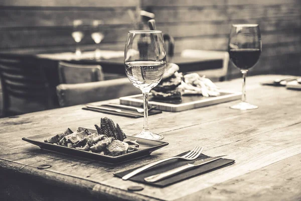 Black and white picture of traditional restaurant atmosphere with tuna tataki and burger with chips in the background on rustic wooden table and glass of white wine