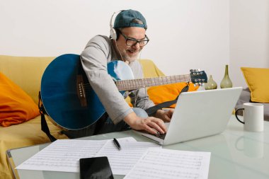 Musician playing the guitar and composing music with his colleagues by video conference through his laptop