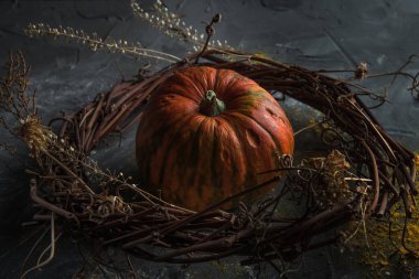 Süslü bir dal çelengi içindeki güzel balkabağı ve karanlık taş bir masanın üzerindeki kuru otlar.