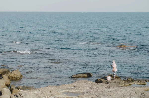 male fisherman in a hat with a fishing rod on the coast of the sea stands on the stone coast. Turkey