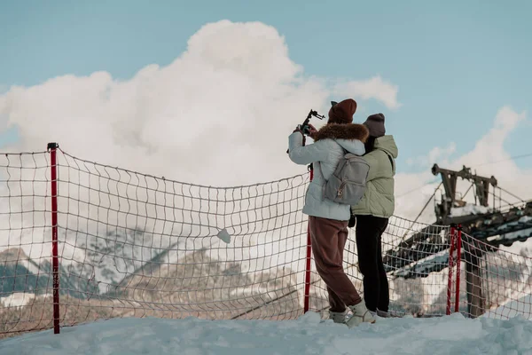 Adler, Rosa Khutor. Gözlem güvertesinde çitin yanındaki dağ manzarasını fotoğraflayan iki kız yürüyüşçü. Arka plan.