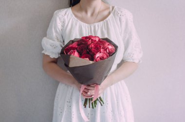 Beyaz elbiseli bir kız kraft kağıdına sarılı bir buket kırmızı gül tutuyor. Görünürde yüz yok