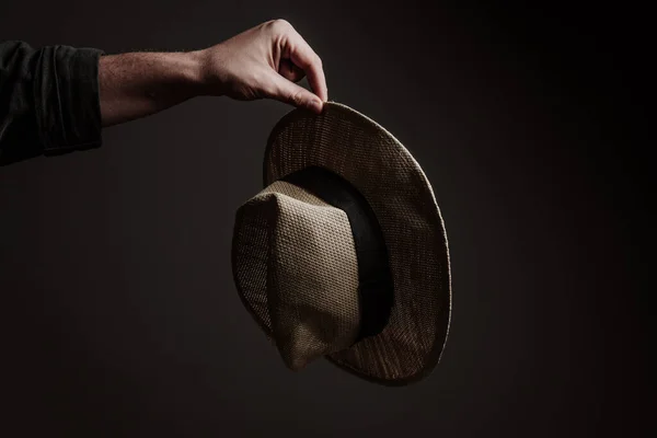 a man\'s hand holding a straw hat on a dark background