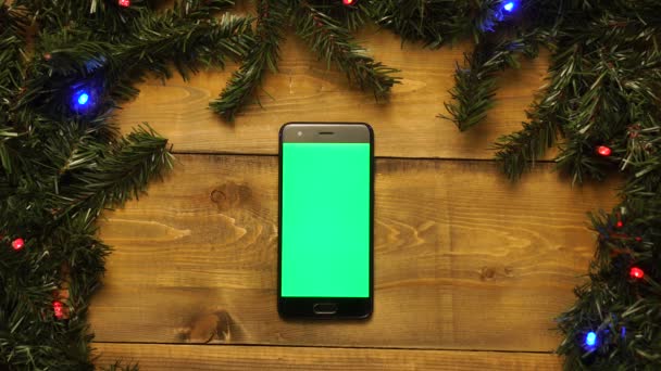 Telefon mit grünem Bildschirm auf dekoriertem Holztisch mit blinkenden Girlanden. — Stockvideo