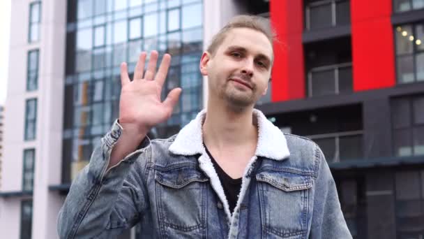 En kjekk mann i jeans jakke som står alene utendørs og hilser ved å vifte med hånden.. – stockvideo