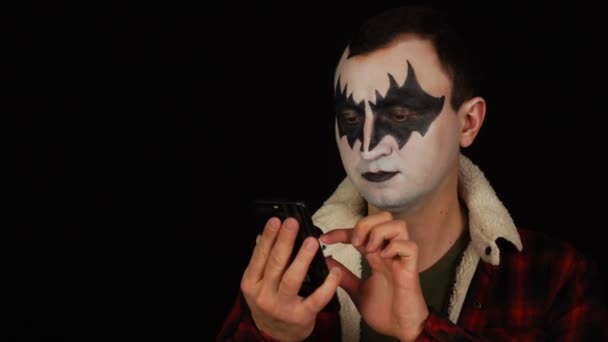 Человек в демоническом макияже, используя свой смартфон на черном фоне — стоковое видео