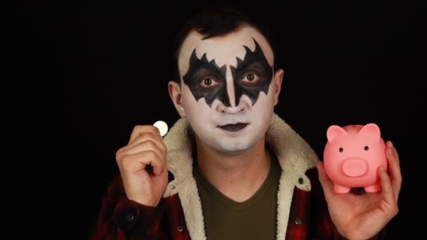 Человек в демоническом макияже кладет монету в копилку и откладывает деньги — стоковое видео