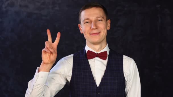 Portret van een stijlvolle jongeman in een vest met vlinderdas en een vrolijk gezicht met vredesgebaar — Stockvideo