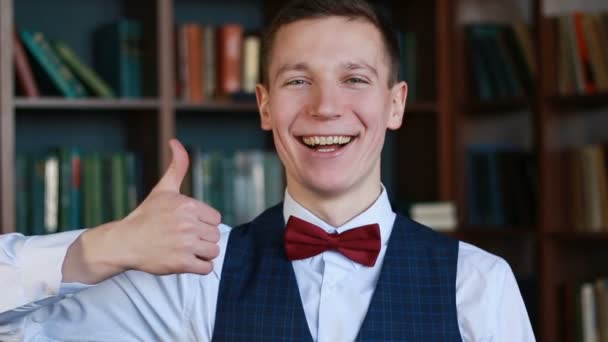 Onnellinen valkoinen mies seisoo yliopiston kirjastossa näyttämässä peukaloa ja hymyilemässä — kuvapankkivideo