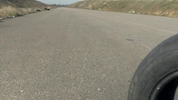 Gamle brugte dæk rullende på asfalten tom vej skubbet af vind og falder hvirvlende. Bane omgivet af gule og grønne bakker . – Stock-video
