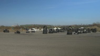 Eski kullanılmış lastikler asfalt yolda yığılmış. Arka planda bir sürü enkaz var. Rüzgar çöp parçalarını hareket ettirir. Parlak güneşli bir gün.