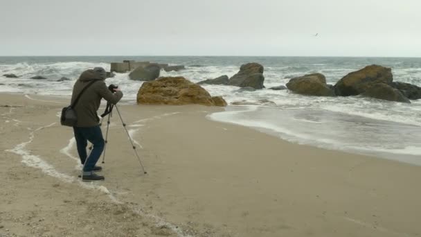 Fotograf upravuje fotoaparát na stativu na písečné pláži blízko skal a bouřce moře. — Stock video