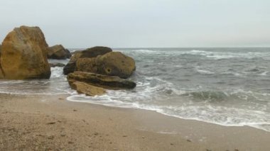 Dalgalı dalgalı deniz, sarı kayalar, gökyüzünü kaplayan yoğun gri bulutlar ile kumlu plaj.
