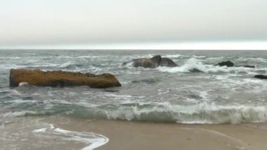 Dalgalı dalgalı deniz, sarı kayalar, gökyüzünü kaplayan yoğun gri bulutlar ile kumlu plaj.