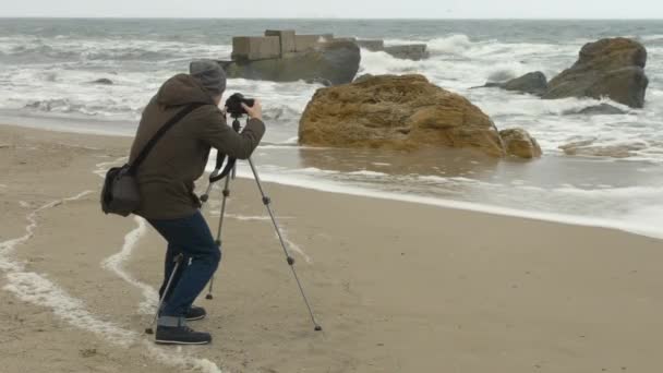 Fotoğrafçı kayalar ve fırtınalı deniz yakınındaki kum plajda tripod kamera ayarlar. — Stok video