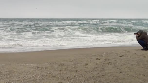 Fotógrafo ajusta la cámara en la playa de arena cerca de las rocas y el mar tormentoso . — Vídeos de Stock