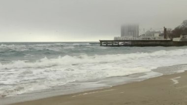 Kumlu plaj ve dalgalı deniz, iskele ve sis, yoğun bulutlar yüksek katlı otel binası