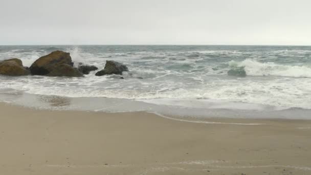 Fotograf dostosowuje kamerę na statywie na piaszczystej plaży w pobliżu skał i burzowego morza. — Wideo stockowe