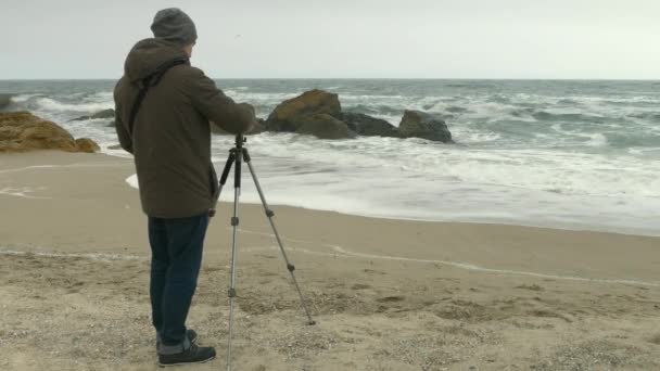 Fotógrafo ajusta la cámara en el trípode en la playa de arena cerca de las rocas y el mar tormentoso . — Vídeos de Stock