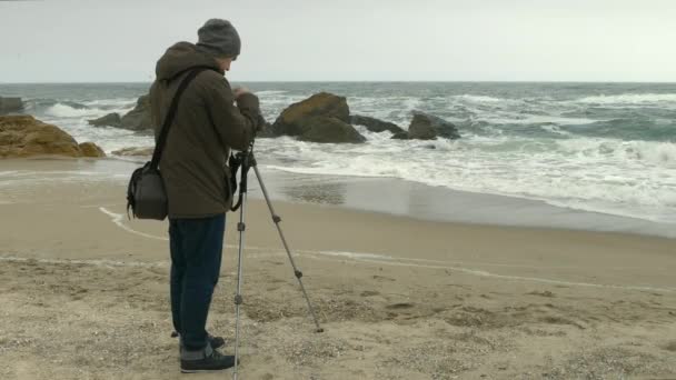Fotograf dostosowuje kamerę na statywie na piaszczystej plaży w pobliżu skał i burzowego morza. — Wideo stockowe