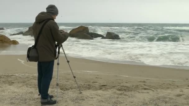 Photographer adjusts camera on tripod on sand beach near rocks and stormy sea. — Stock Video