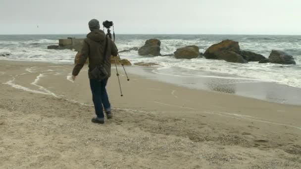 Fotógrafo ajusta câmera em tripé na praia de areia perto de rochas e mar tempestuoso . — Vídeo de Stock