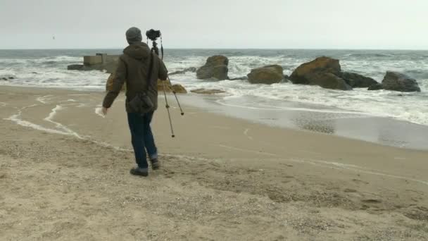 Fotograf stellt Kamera auf Stativ am Sandstrand in der Nähe von Felsen und stürmischer See ein. — Stockvideo