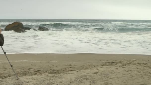 Photographer adjusts camera on tripod on sand beach near rocks and stormy sea. — Stock Video