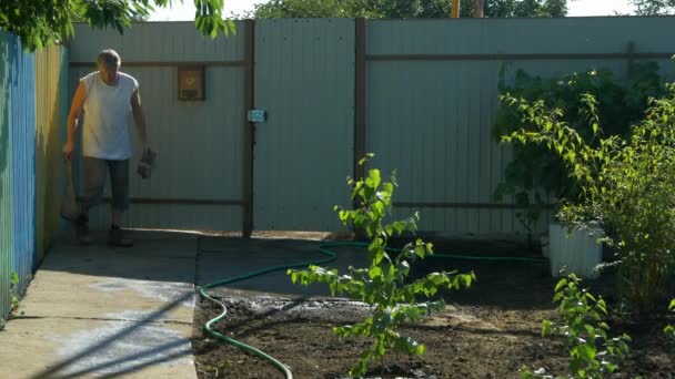 Gardener sweeping concrete walkway in garden after watering trees and plants. — Stock Video