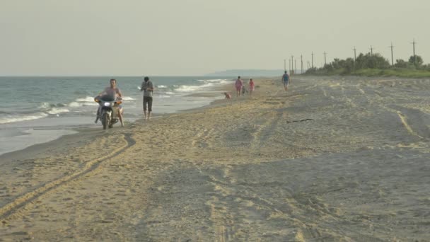 Um homem monta uma moto em uma praia de areia selvagem perto da água do mar . — Vídeo de Stock