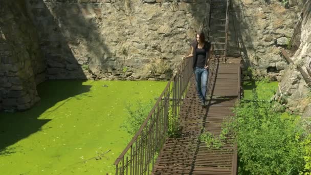 Young beautiful girl walks on thin ruined rusty metal bridge over green water. — Stock Video