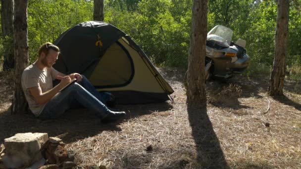 Μοτοσικλετιστής κάθεται κοντά σε σκηνή στο δάσος και να γράφει ένα βιβλίο στις διακοπές κοντά στο στρατόπεδο — Αρχείο Βίντεο
