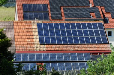 A close-up of multiple solar panel arrays on the tiled roof of a building. The panels vary in age and type, indicating different installations over time. The surrounding foliage is lush and green.