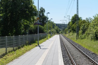 A view down an empty, sunlit train platform and adjacent railway track. The platform is paved with light-colored tiles, and a sign and streetlights are visible. Lush green trees and vegetation line