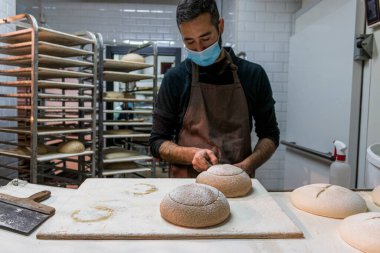 Yüz maskeli beyaz adam arka planda çiğ ekmek ve fırın arabaları kesiyor.