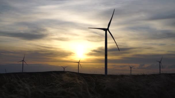 Movimiento de las palas de un gran aerogenerador en un campo sobre un fondo de naranja puesta de sol en el horizonte con una hermosa colinas. Fuentes de energía alternativas. Parque de viento. Energía ecológica.Industrial — Vídeos de Stock