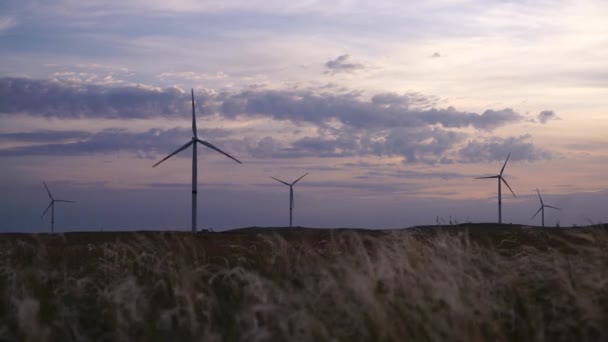 Poruszaj ostrzami dużej turbiny wiatrowej w polu na tle pomarańczowego zachodu słońca na horyzoncie z pięknymi wzgórzami. Alternatywne źródła energii. Park wiatrów. Energia ekologiczna.Przemysł — Wideo stockowe