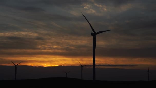 Движение лопастей большой ветряной турбины в поле на фоне оранжевого заката на горизонте с красивыми холмами. Альтернативные источники энергии. Ветреный парк. Экологическая энергетика. — стоковое видео